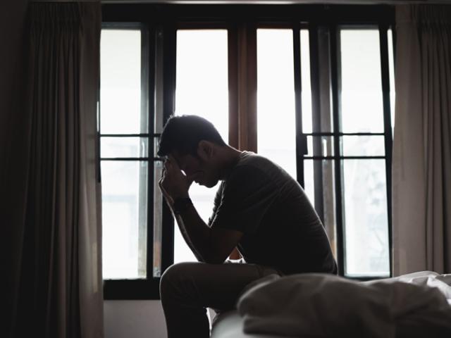 Hombre sentado en la cama con la cabeza entre las manos, expresando tristeza o malestar emocional.