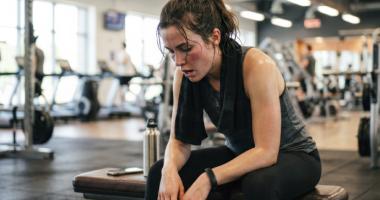 Mujer agotada sentada en el gimnasio, tras un entrenamiento intenso, evidenciando fatiga y posibles agujetas.  